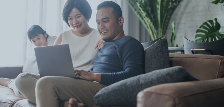 A family of mother and father with daughter on a sofa looking at a laptop.